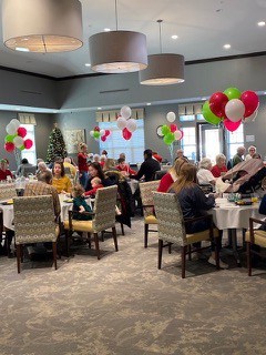 Groups of people celebrating Christmas sit around tables with red and green balloons in the dining room. Grey walls and brown patterned carpet