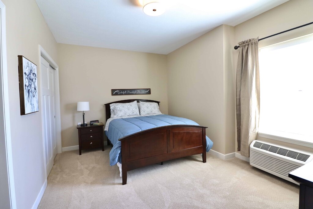 model bedroom of a Traditions at North Bend unit featuring a bed, a night stand a closet door, a window and carpeted floor
