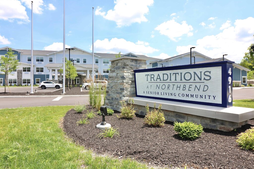 an entrance sign for Traditions at North Bend sitting in a pile of mulch in front of a blue and white senior living building