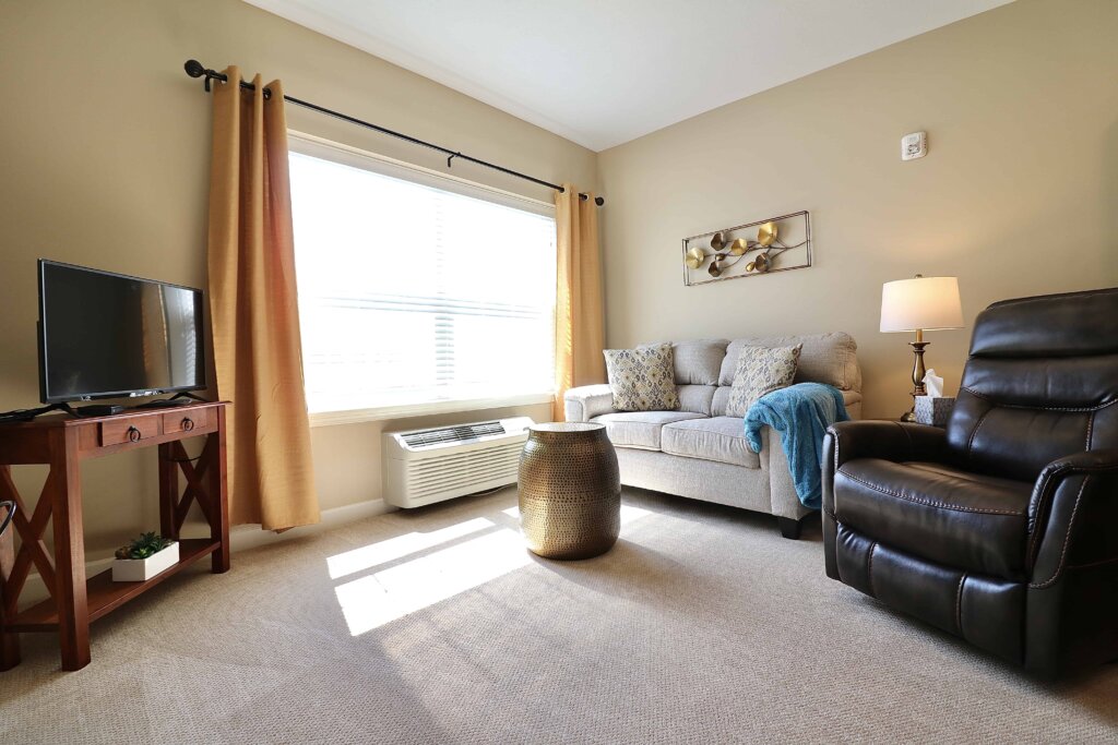 model living room in a Traditions at North Bend unit featuring a large window, tv, small couch and reclining chair
