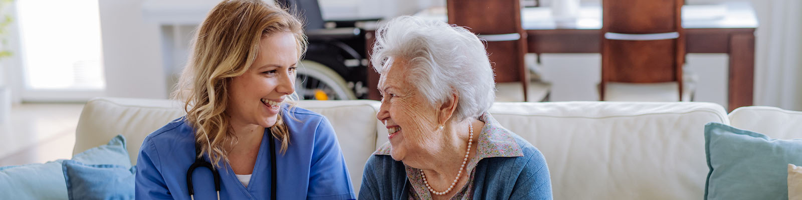 A senior woman sitting on a couch with her nursing assistant as they smile at each other