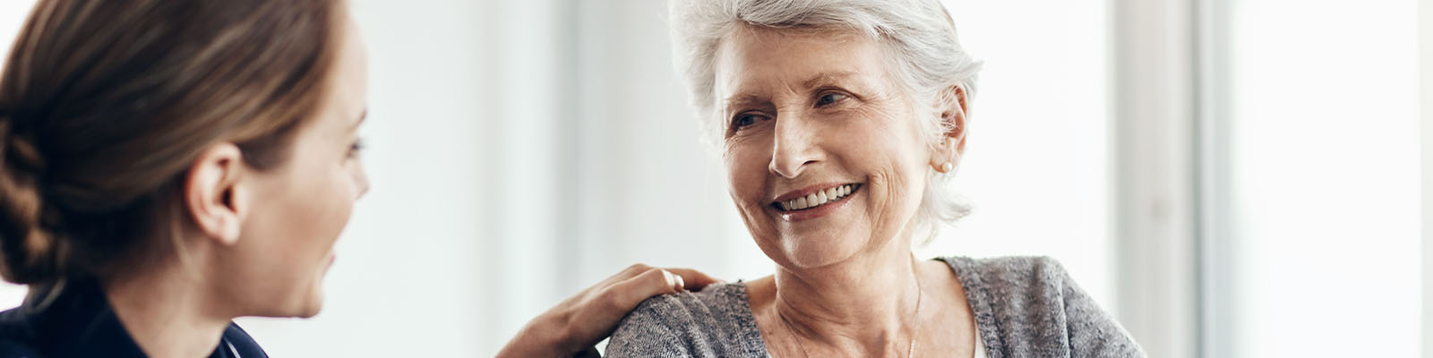 A senior woman sitting in a wheelchair looking at her nursing assistant kneeling down beside her