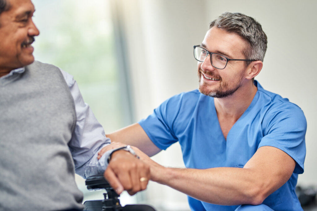 Male nurse squatting next to male memory care resident chatting and smiling at Traditions at North Bend in Cincinnati, OH.