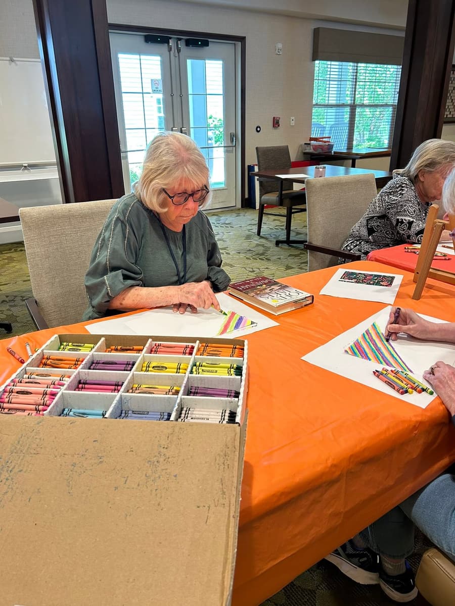 Two residents focus on a crayon coloring activity at a table in the common area at Traditions at North Bend, with a large box of crayons shared between them.