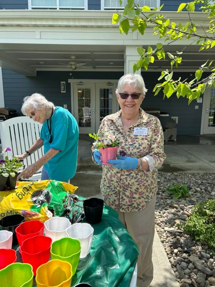 Two residents participate in an outdoor potting activity at Traditions at North Bend, with one smiling and holding a pink pot of freshly planted flowers.